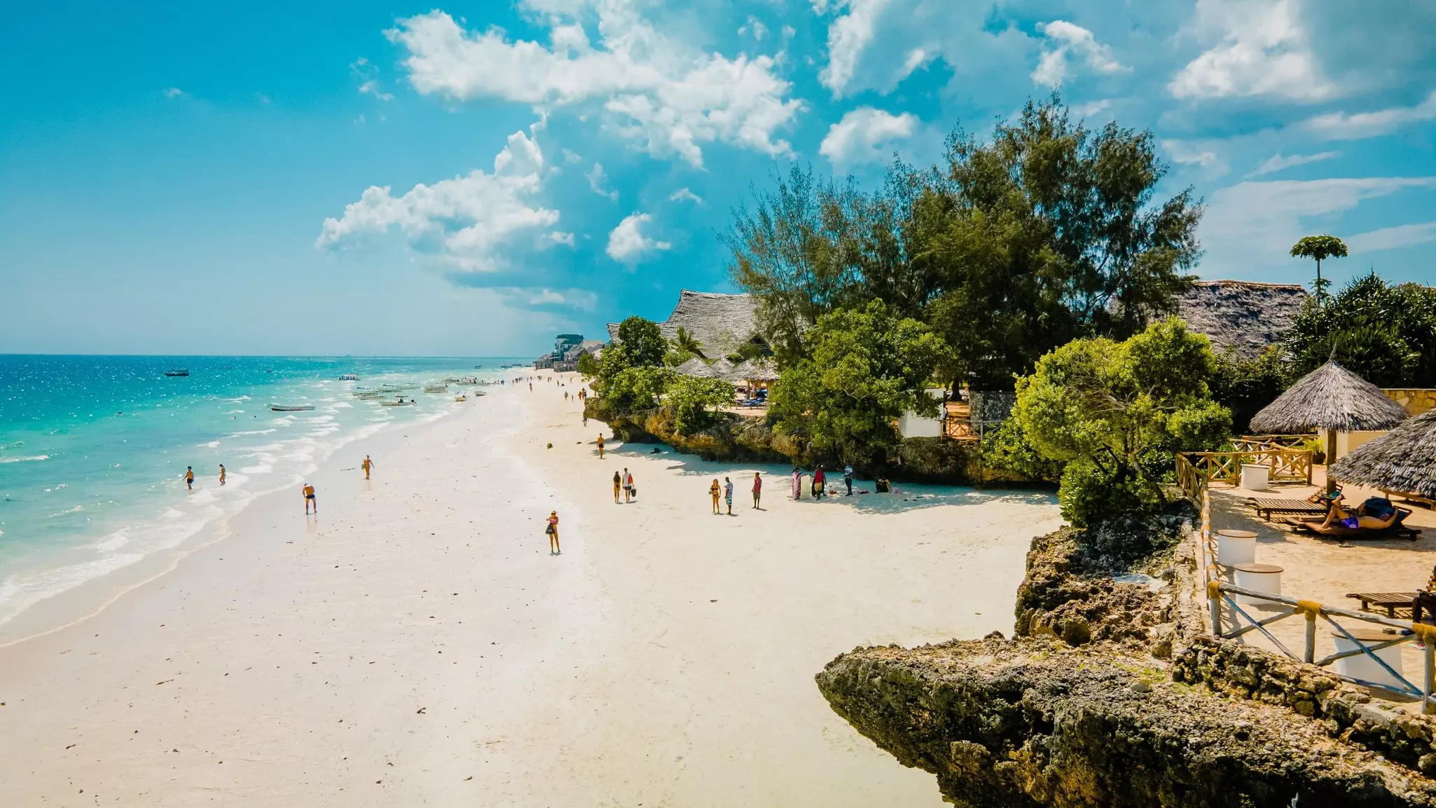 A wide shot of a white-sand beach with people walking and swimming, and thatched-roof houses and trees visible on shore.