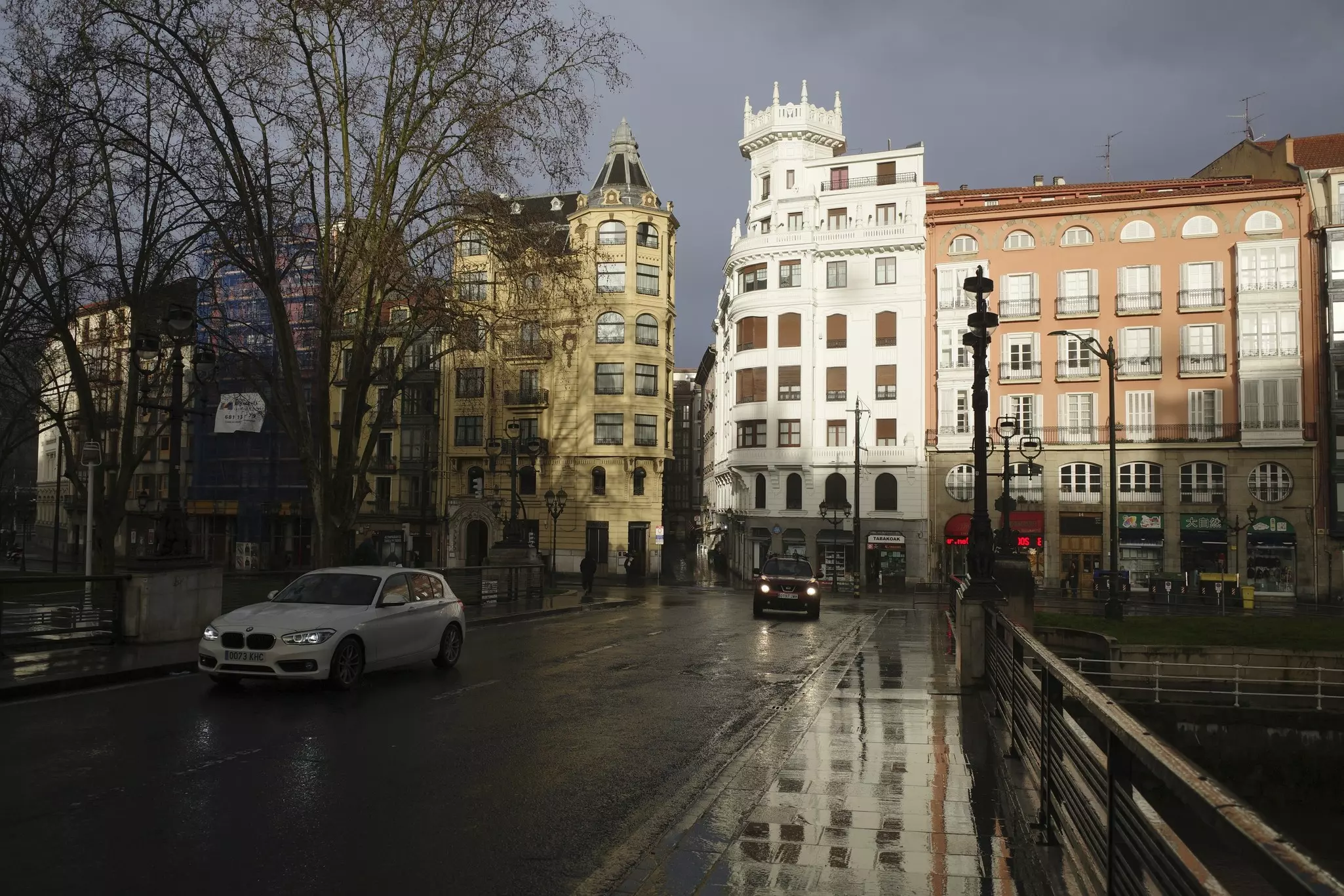 Cars on a city street whose surface is wet with rain. Storm clouds are overhead, while sunlight dramatically illuminates buildings along the street.