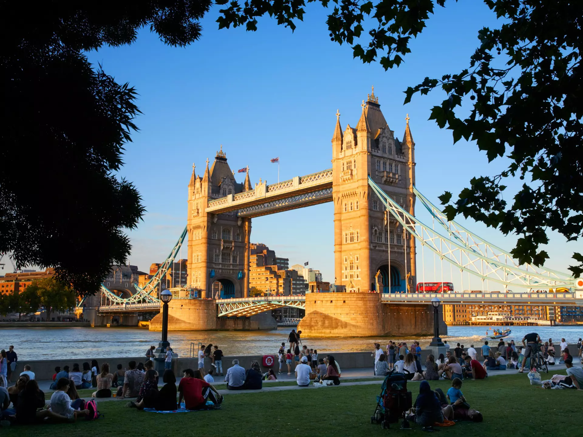 People sit on grass near a Gothic-style bridge with two towers on a summer's day