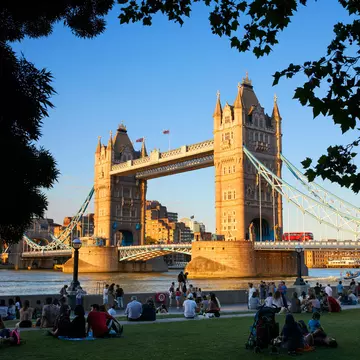 People sit on grass near a Gothic-style bridge with two towers on a summer's day