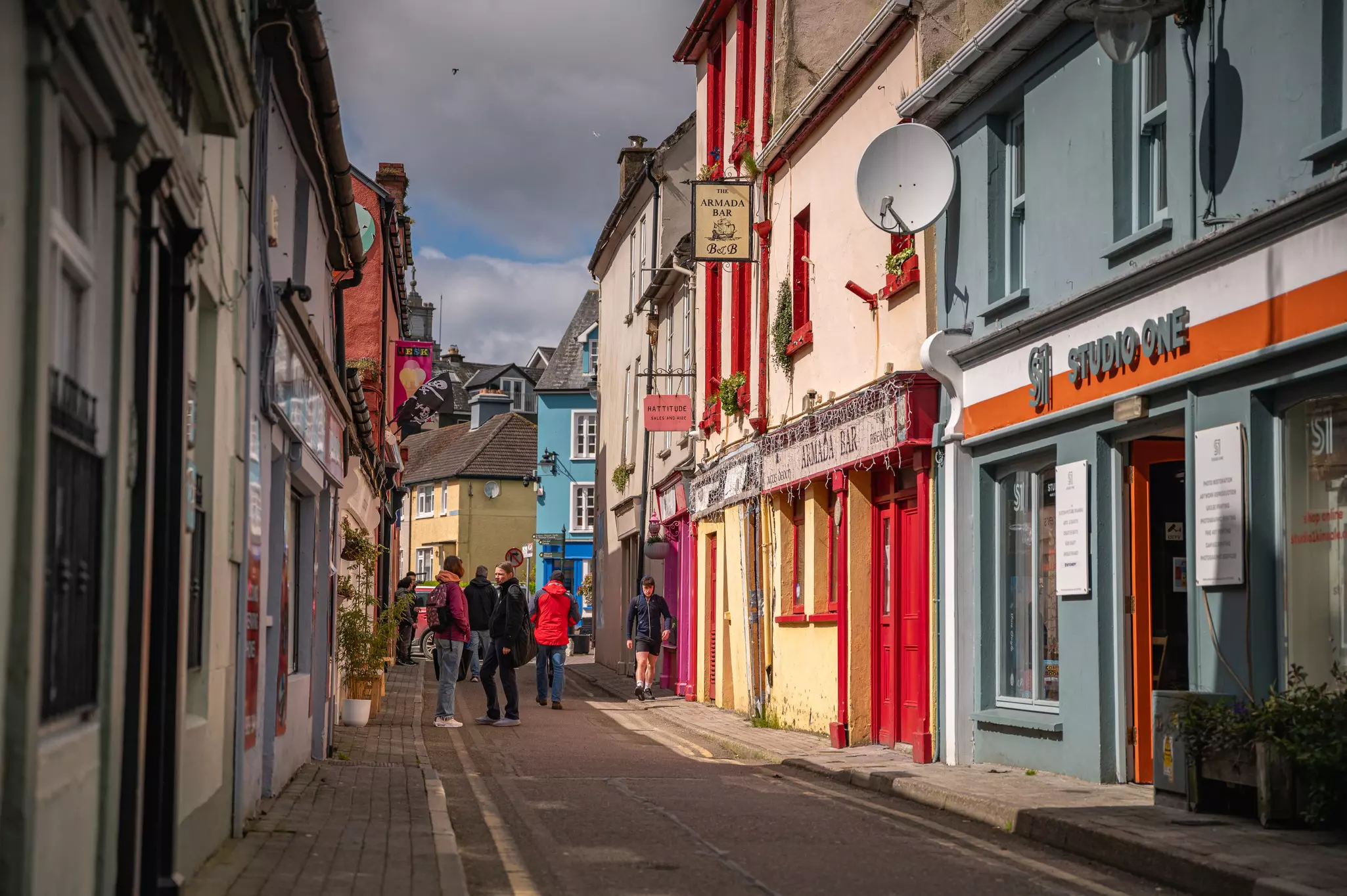 A narrow main street of a small village. People in colorful coats are standing on the street chatting. The street is lined with pubs and restaurants.