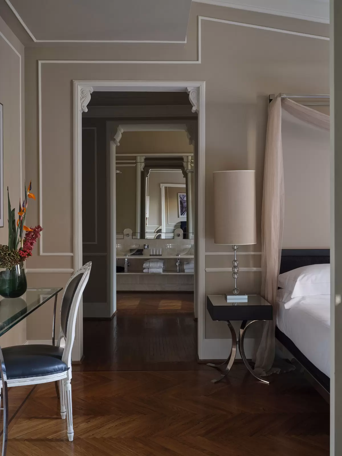 View of a hotel guest room with the edge of a four-posted bed, and a doorway looking into a double-sink bathroom