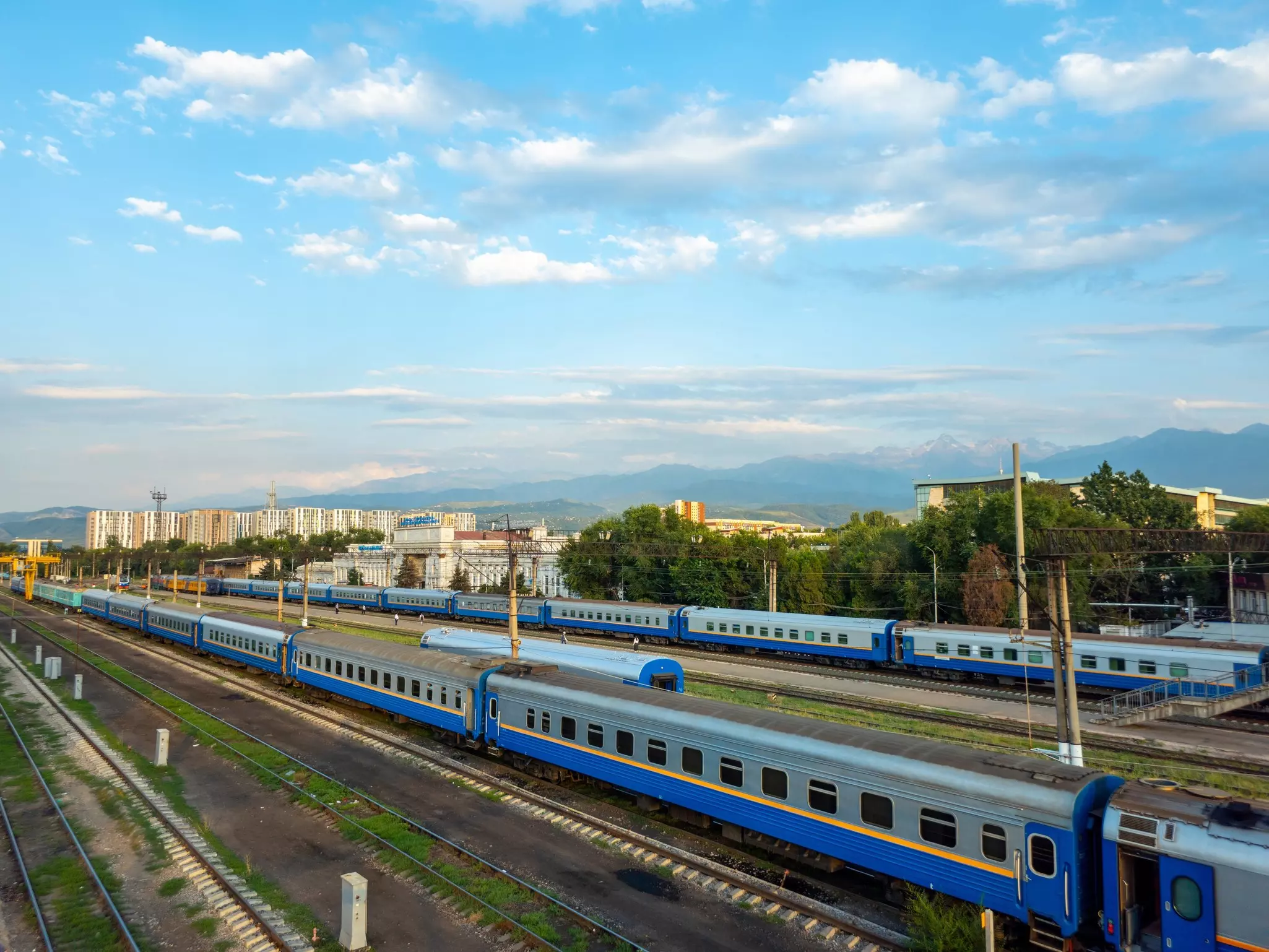 Blue and gray trains with a yellow stripe down the length of the carriages wait on rails at the edge of a city station.