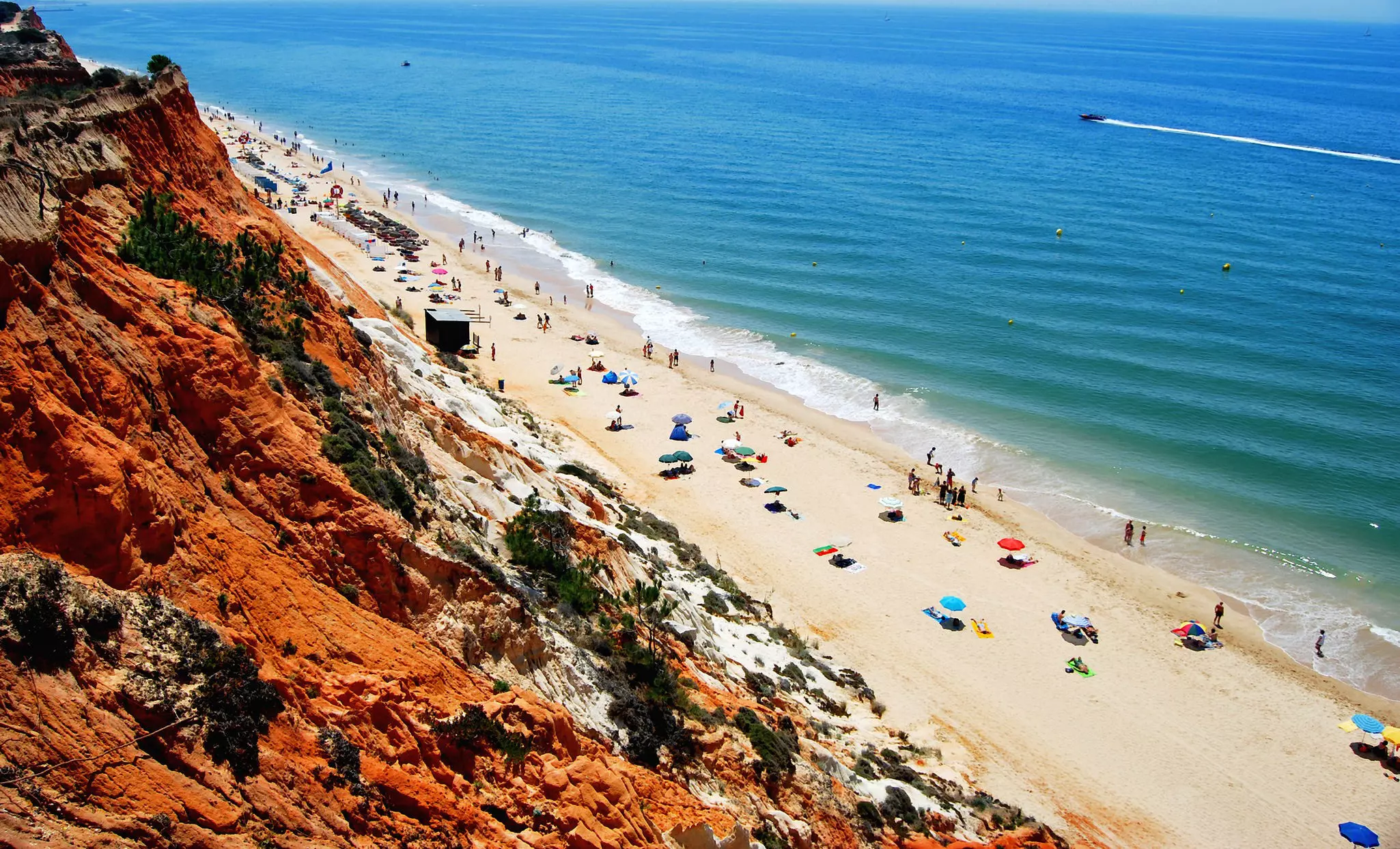 Bird's-eye view of a long, sandy beach with umbrellas, people and the sea beyond. A red cliff is in the foreground.