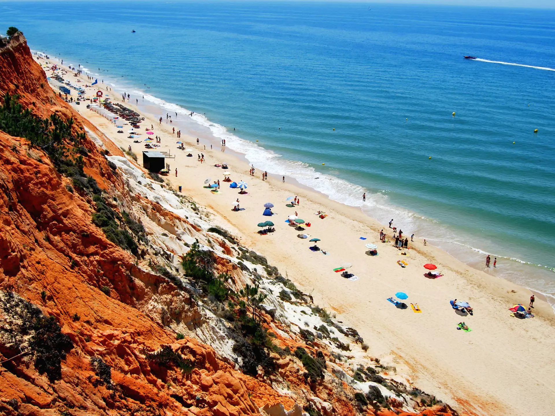 Bird's-eye view of a long, sandy beach with umbrellas, people and the sea beyond. A red cliff is in the foreground.