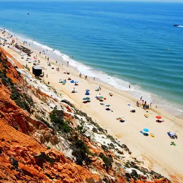 Bird's-eye view of a long, sandy beach with umbrellas, people and the sea beyond. A red cliff is in the foreground.