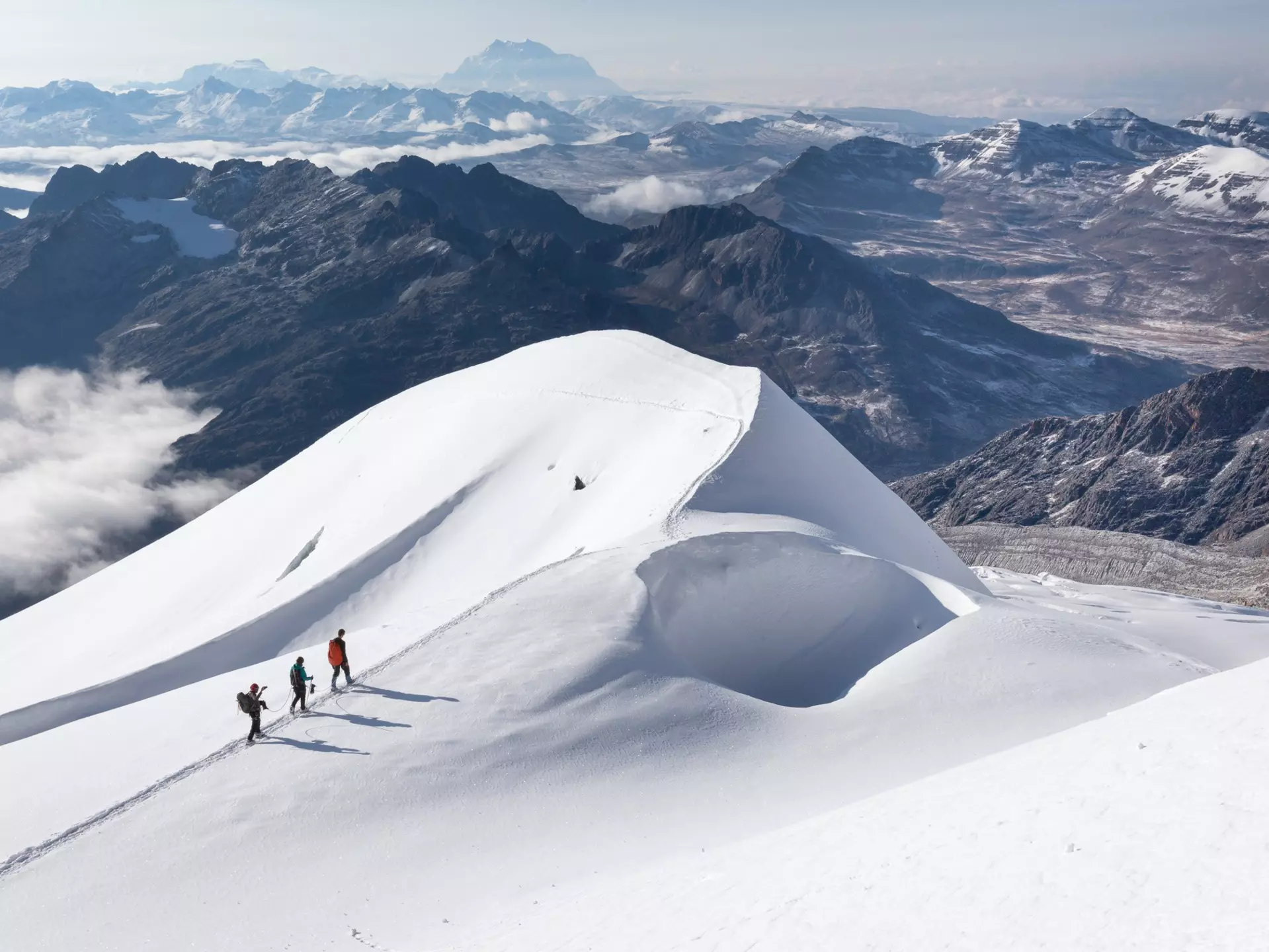 Three mountaineers on a snow ridge with cloud-covered mountains below, near or on Huayna Potosí