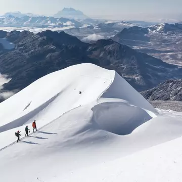 Three mountaineers on a snow ridge with cloud-covered mountains below, near or on Huayna Potosí