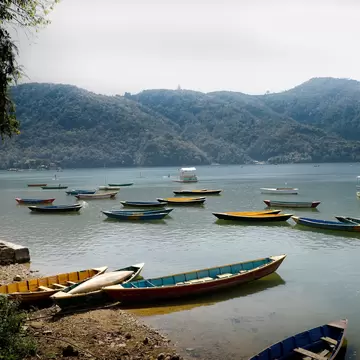 Painted wooden boats rest at a pebbly shoreline and float on calm grey-green water beneath hazy wooded hills
