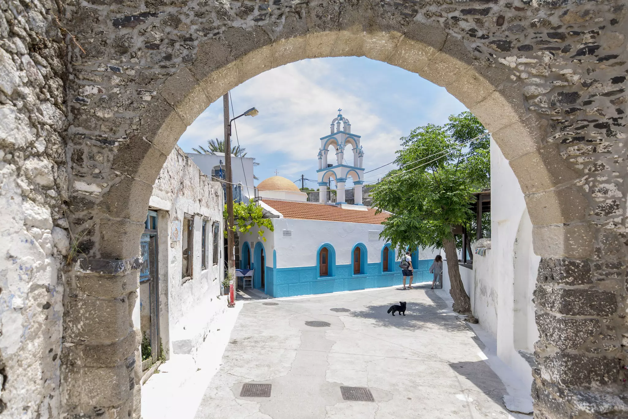 Whitewashed buildings, a small four-legged creature and a couple are viewed through a stone arch.