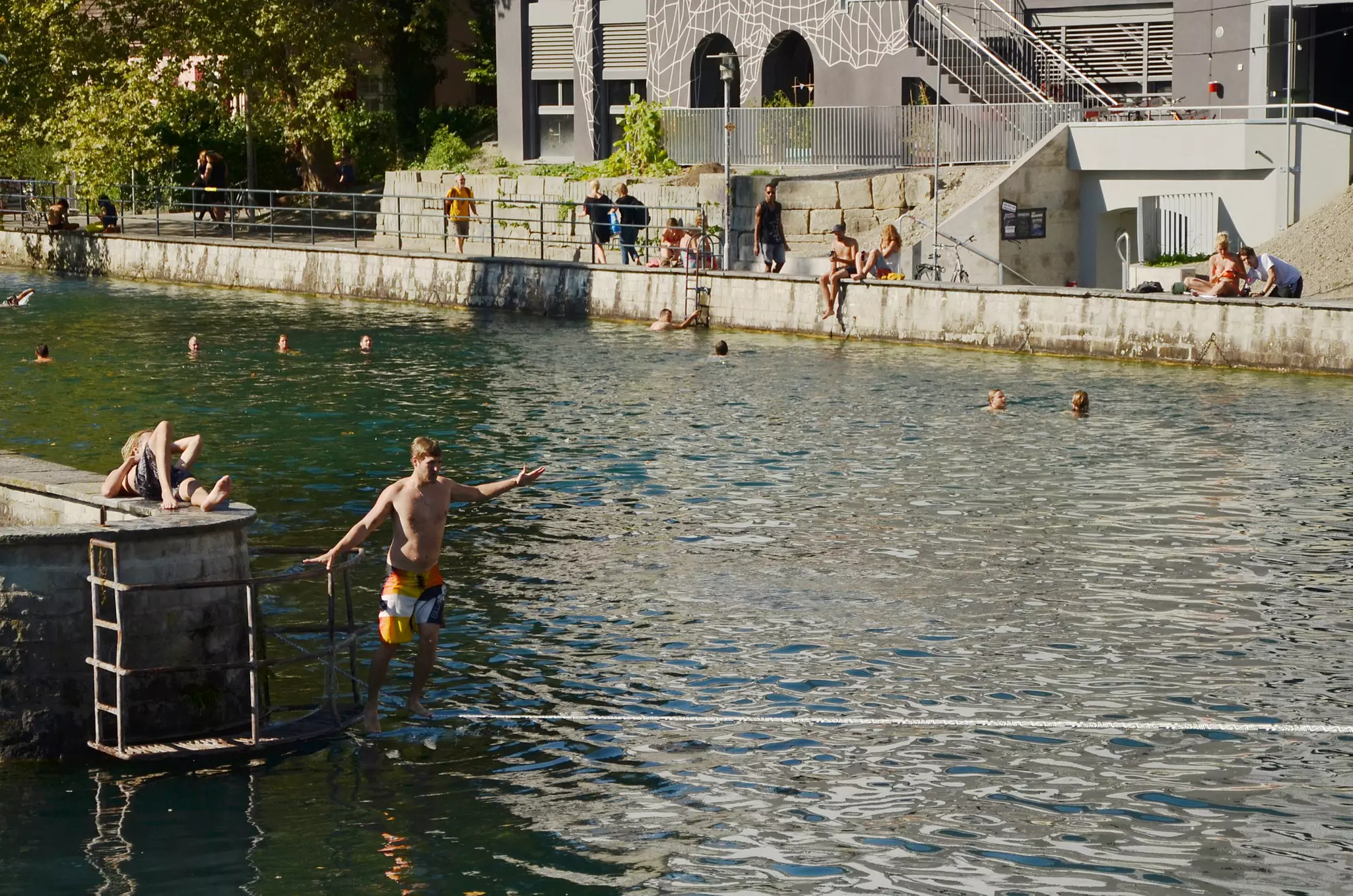 A young man wearing a bathing suit walks on a tightrope over a river in a city. Other people are in swimming the water, and along the banks of the river in the sun.