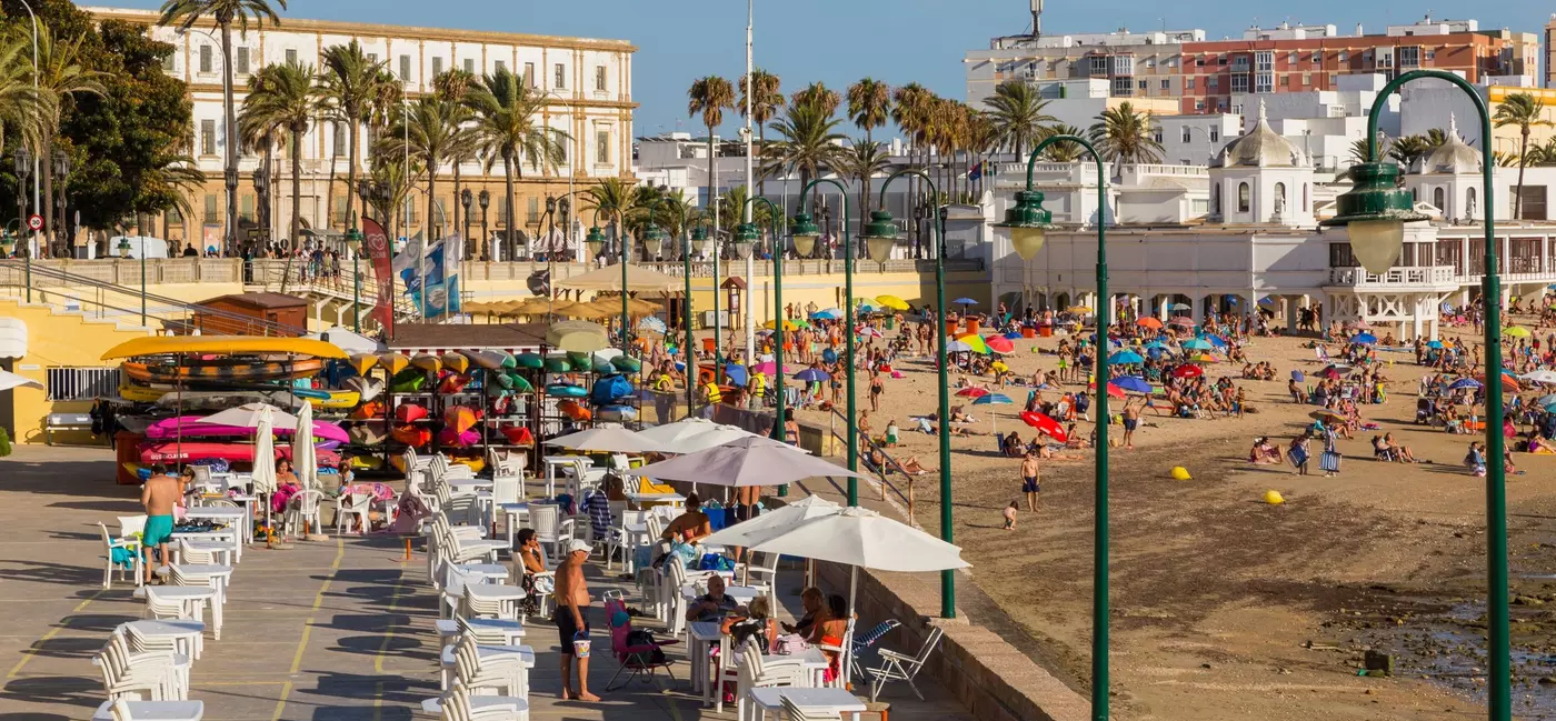 People gather at white plastic tables under white umbrellas beside a beach that's filled with people sitting under colorful parasols.
