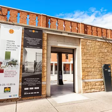 A sandstone and red-brick wall with a deep doorway and two tall exhibition banners in Greek and English hanging beside it.