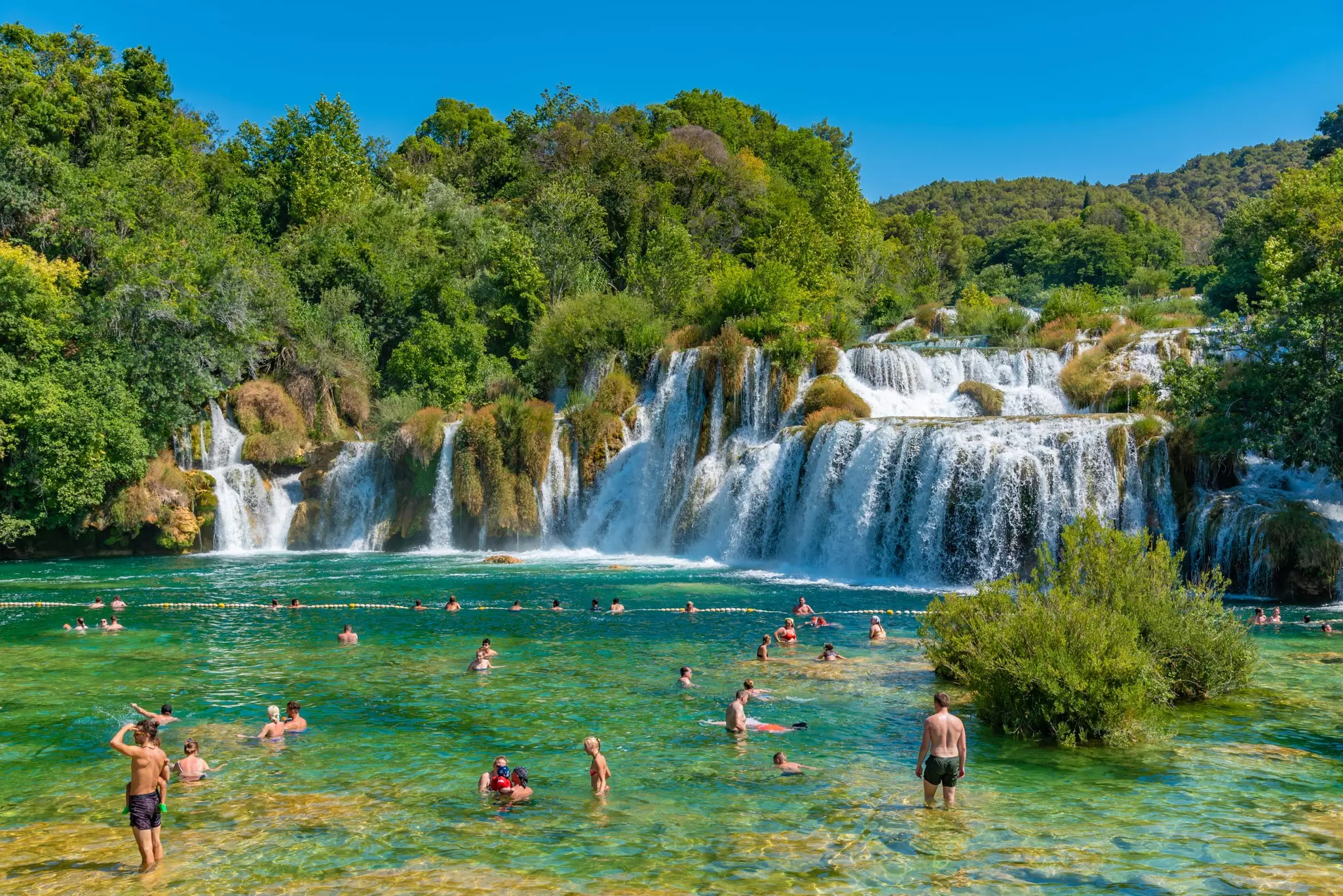 People swimming in a river by a waterfall surrounded by green trees on a sunny day