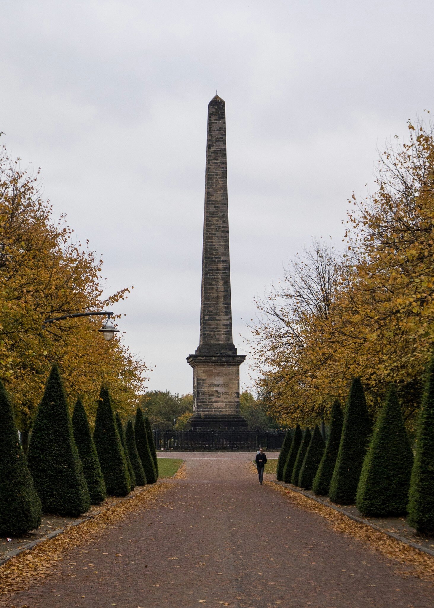 A man walks on a leaf-covered path with the Nelson monument in the background in the Glasgow Green during autumn.