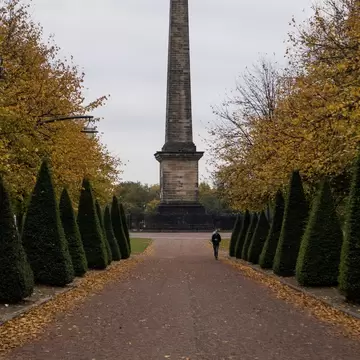 A man walks on a leaf-covered path with the Nelson monument in the background in the Glasgow Green during autumn.