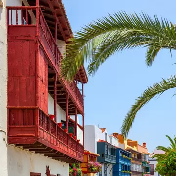 A red wooden balcony and a row of blue, green and yellow balconied houses on a sunny street in Santa Cruz de La Palma.