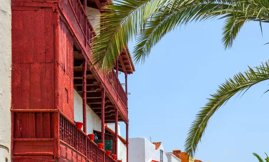 A red wooden balcony and a row of blue, green and yellow balconied houses on a sunny street in Santa Cruz de La Palma.