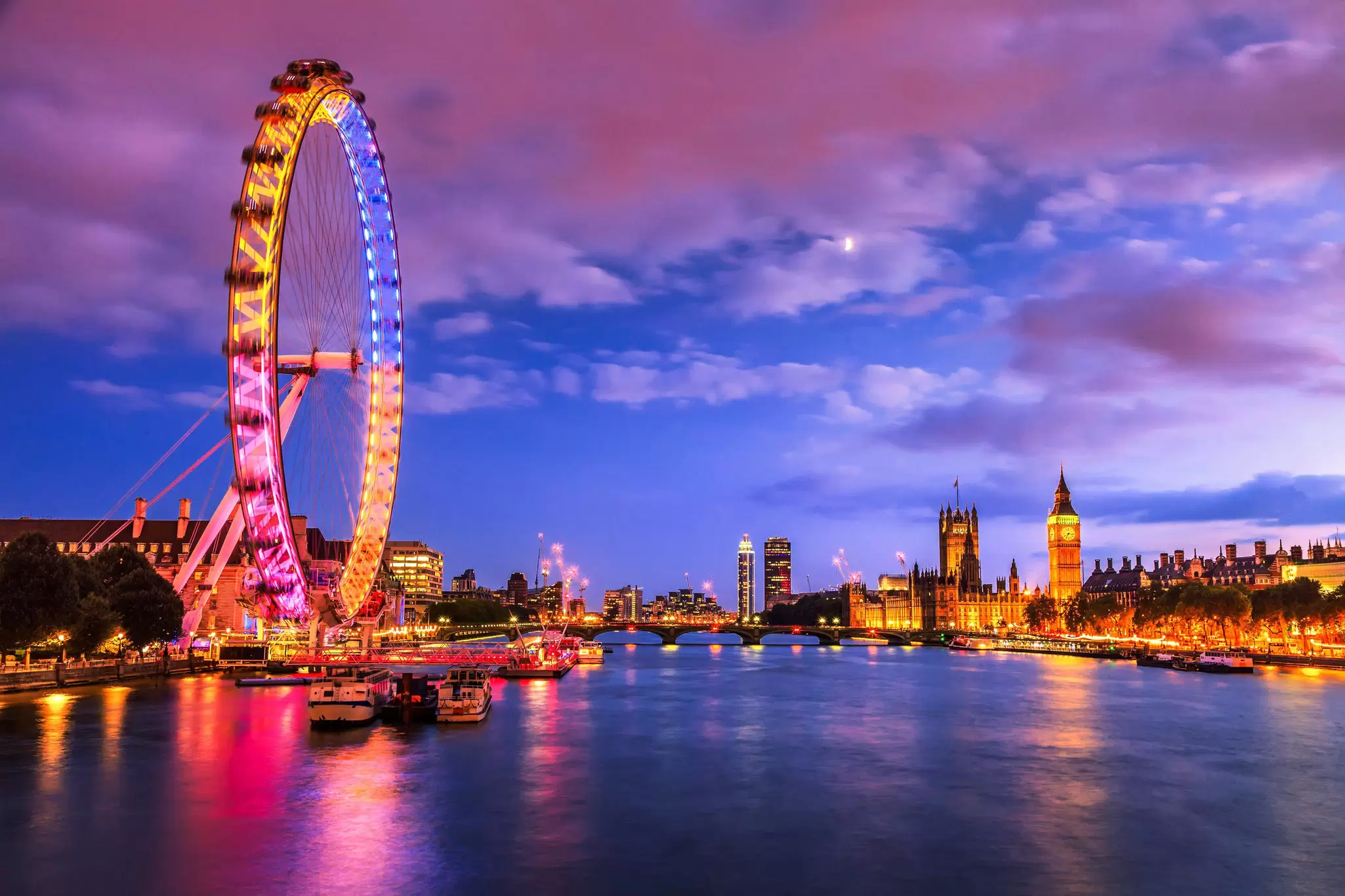 A night time shot of a river with a large Ferris wheel lit up in pink, yellow and blue to the left, and a huge neo-Gothic building to the right