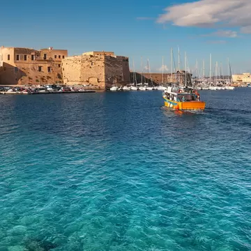 An ancient coastal city seen from sea, with a fortified castle, yacht marina and fishing boat on sunny day.