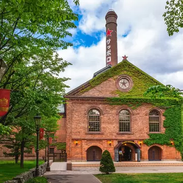 A red-brick building partly covered in ivy, with a tall chimney marked with a red star, behind trees and lampposts.