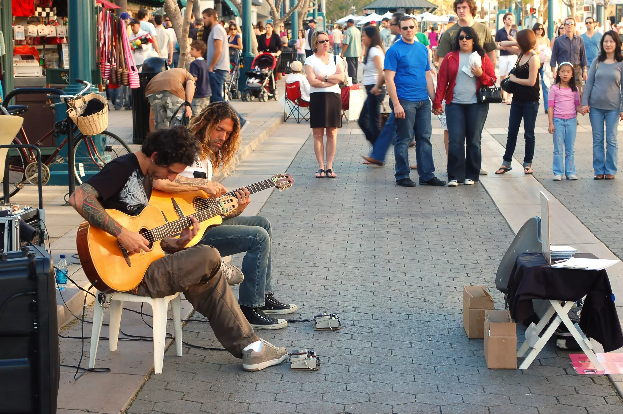 Two guitarists busk in a pedestrianized zone with plenty of onlookers
