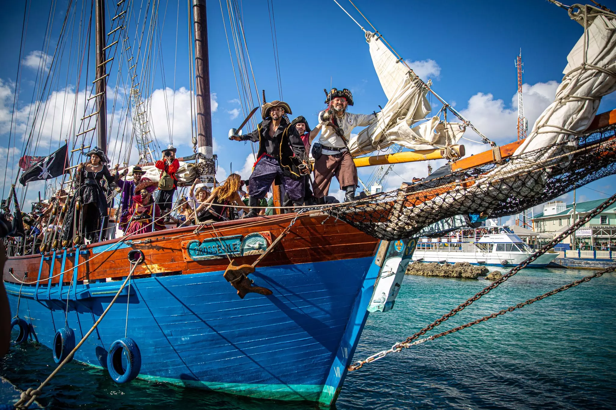 People dressed as pirates hold cutlasses and pose as they head out on an old-fashioned ship flying a skull and crossbones flag.