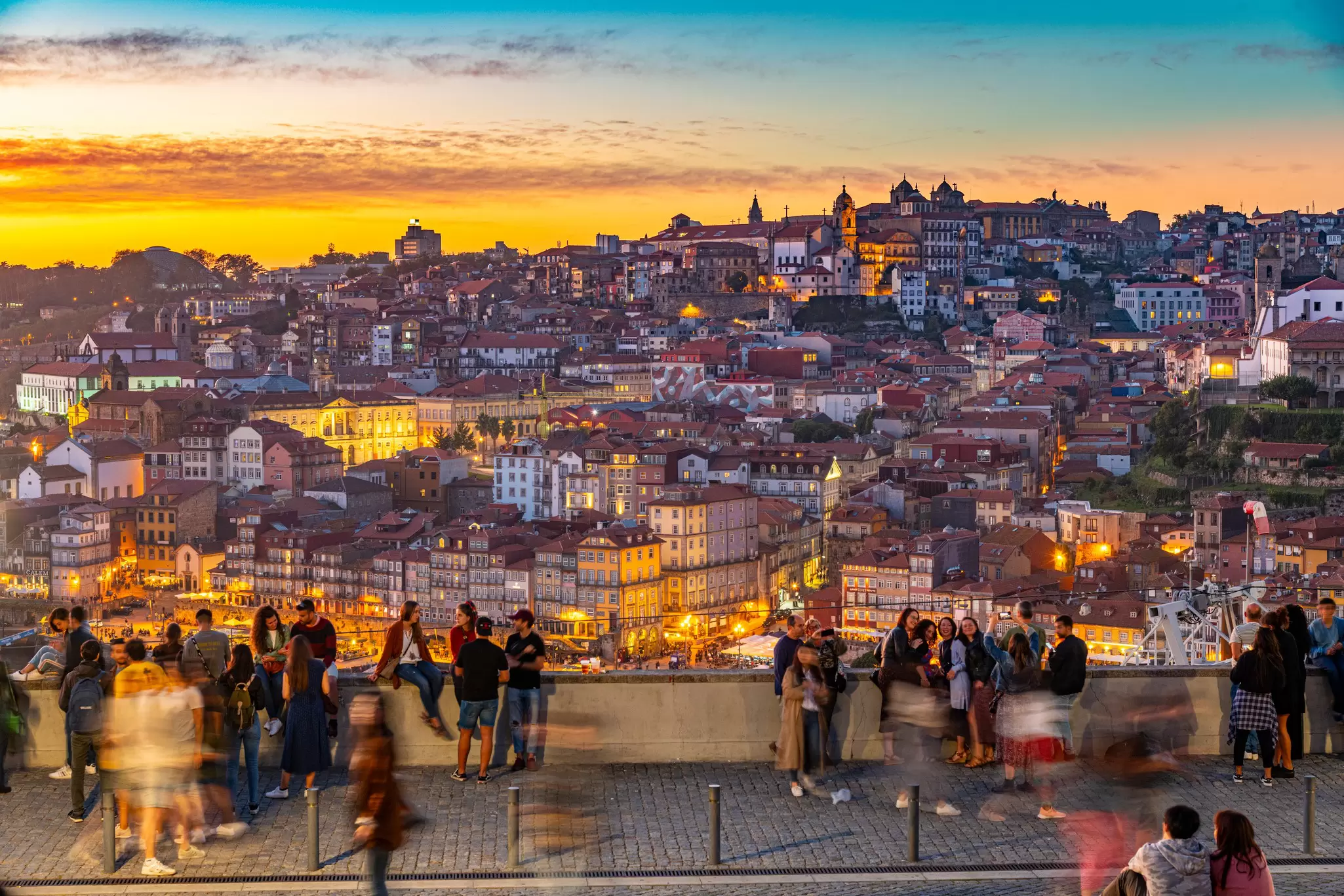 People gather at an overlook over a city lit up below just after sunset.