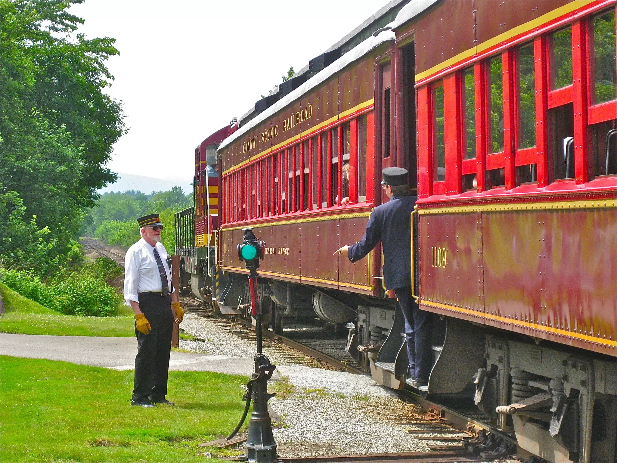 A uniformed conductor stands next to a historic train, speaking with another conductor on the train.
