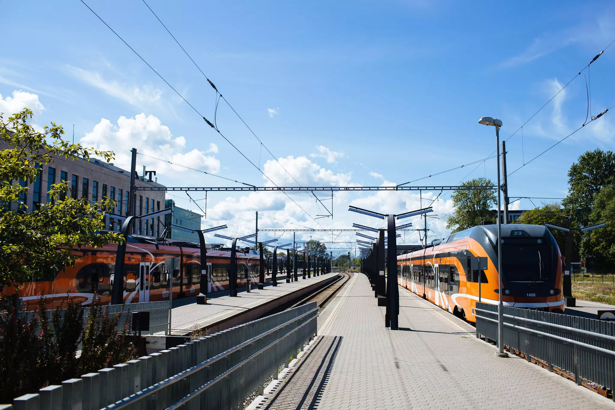 Wide shot of outdoor rail station with yellow train on right tracks and red train on left, with stone platform in between on a sunny day.
