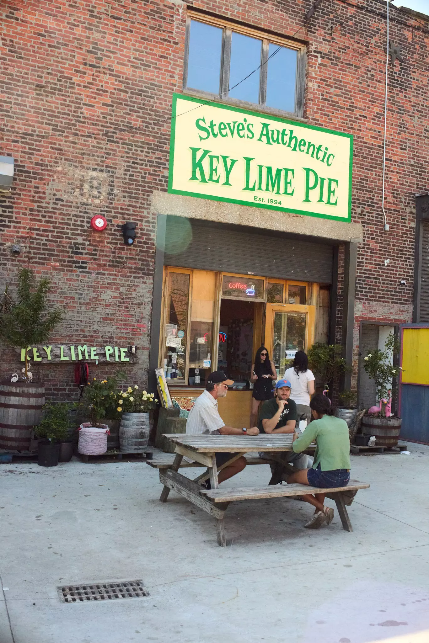 People sit at picnic tables outside a redbrick building with a green sign for Steve's Authentic Key Lime Pie.