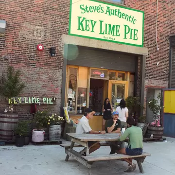 People sit at picnic tables outside a redbrick building with a green sign for Steve's Authentic Key Lime Pie.