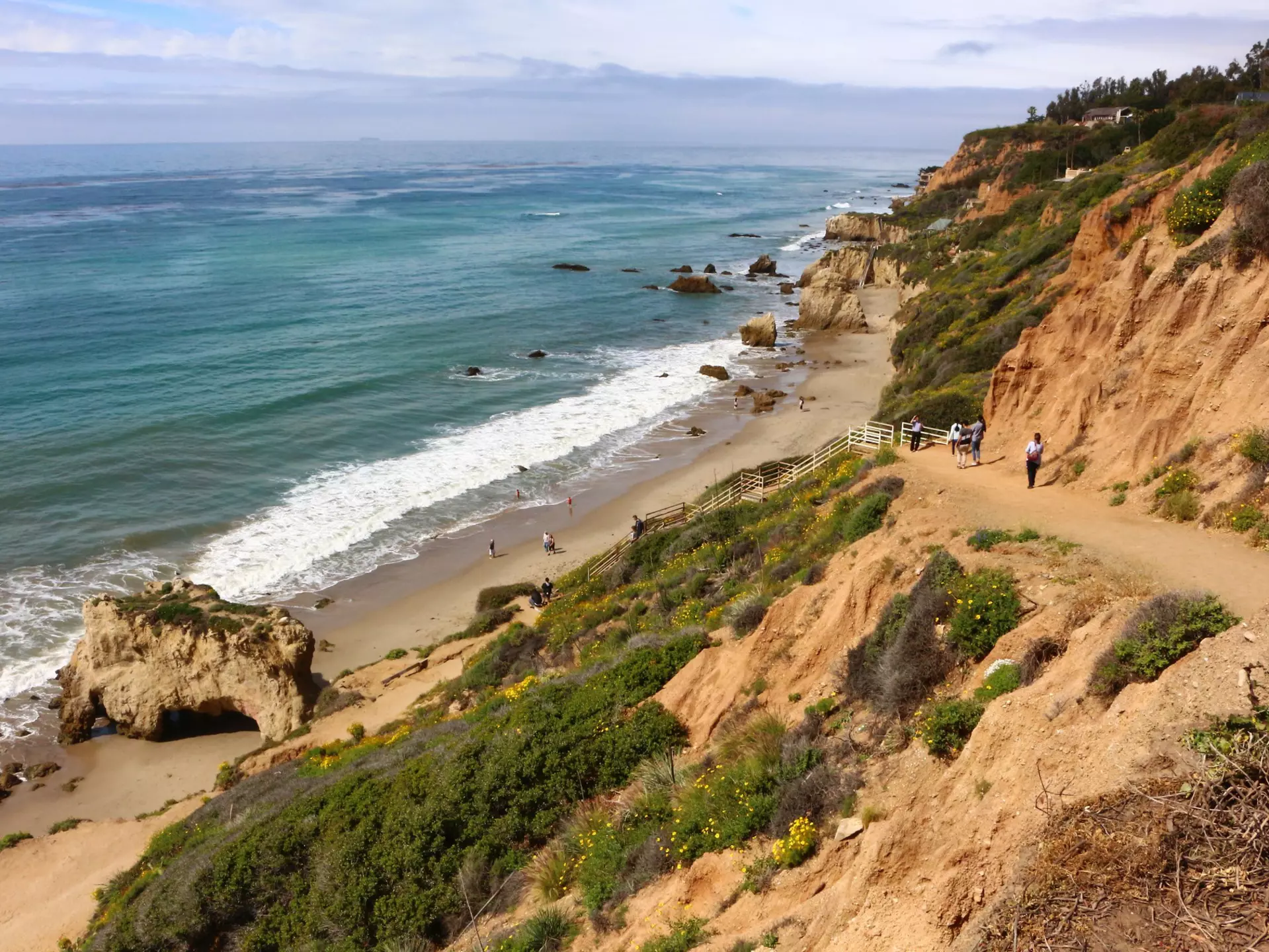 People walk down a sandy coastal path towards a beach with several rock stacks and arches.