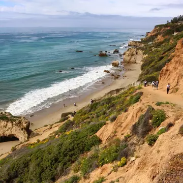 People walk down a sandy coastal path towards a beach with several rock stacks and arches.