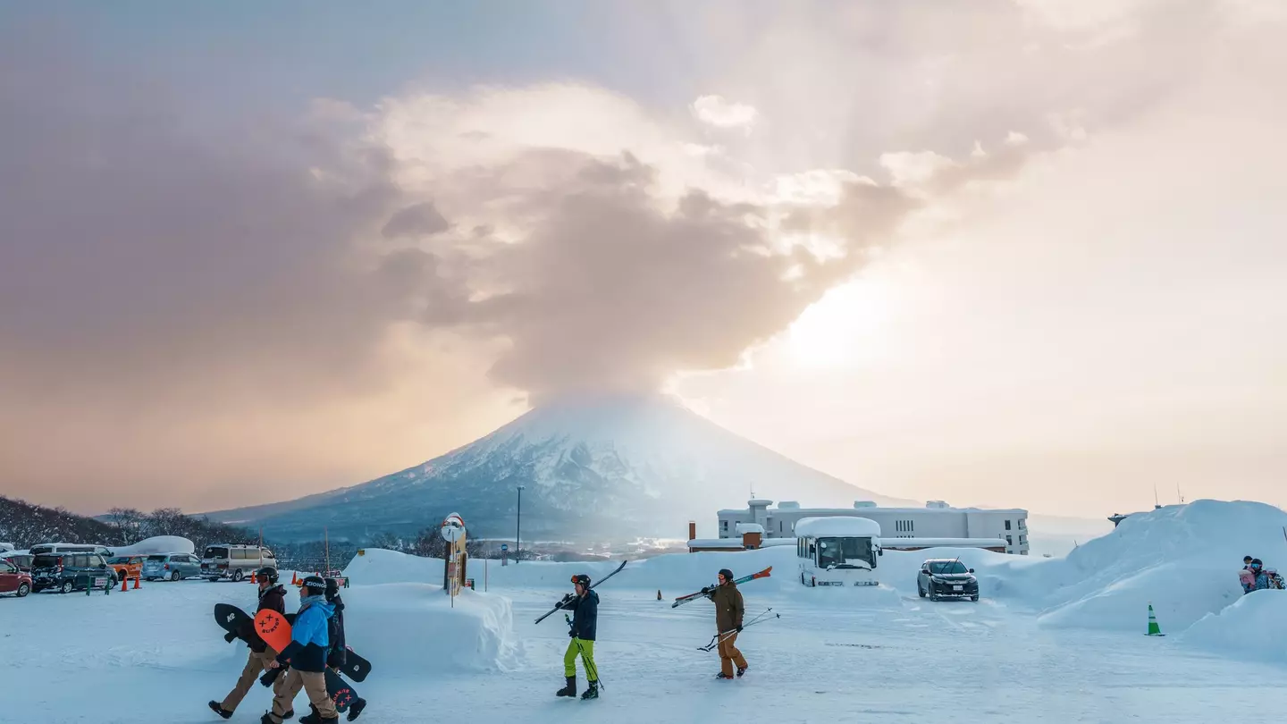 People carry their skis and snowboards at a snowy mountain resort. A huge stratovolcano is visible in the distance.