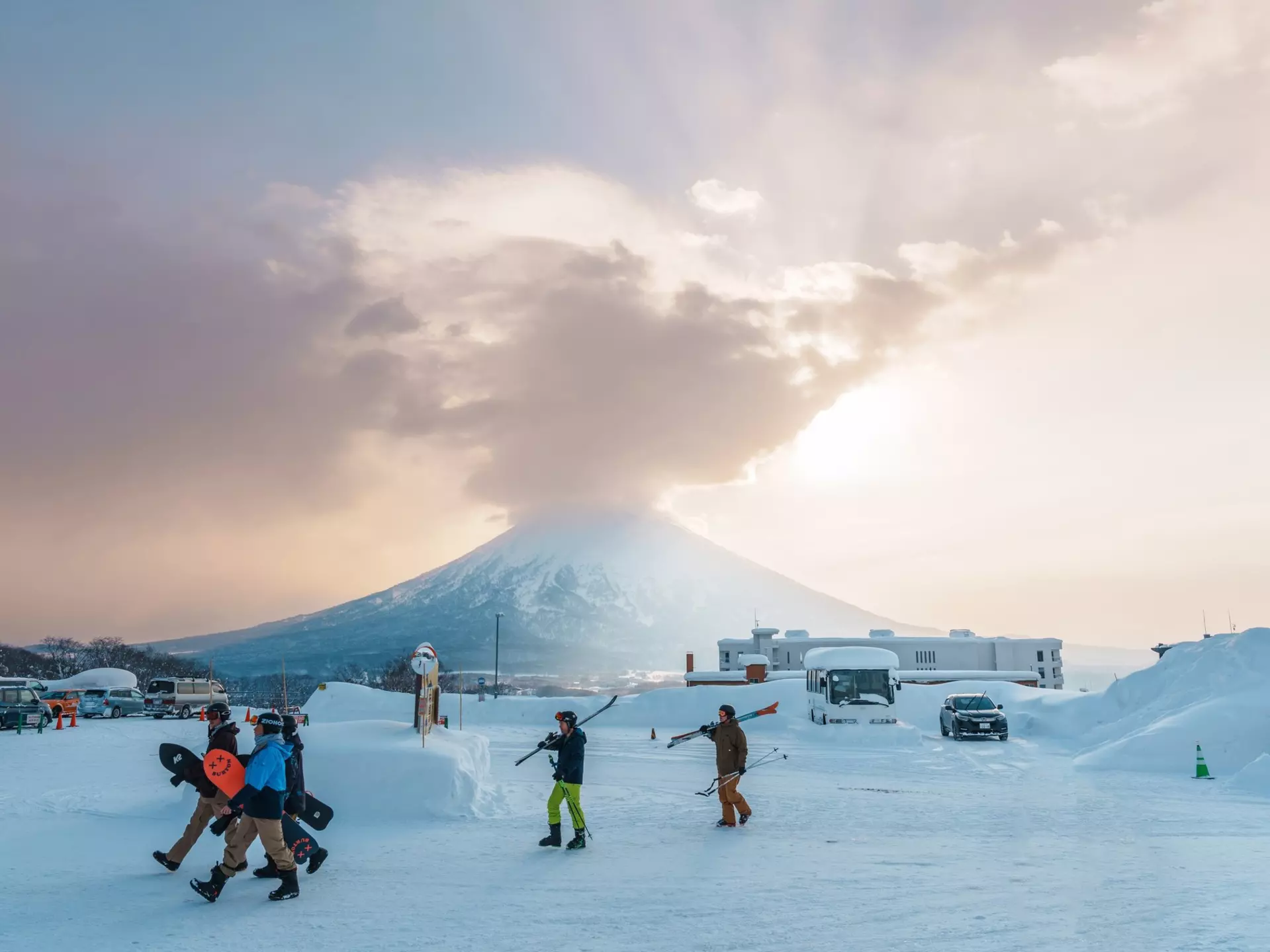 People carry their skis and snowboards at a snowy mountain resort. A huge stratovolcano is visible in the distance.