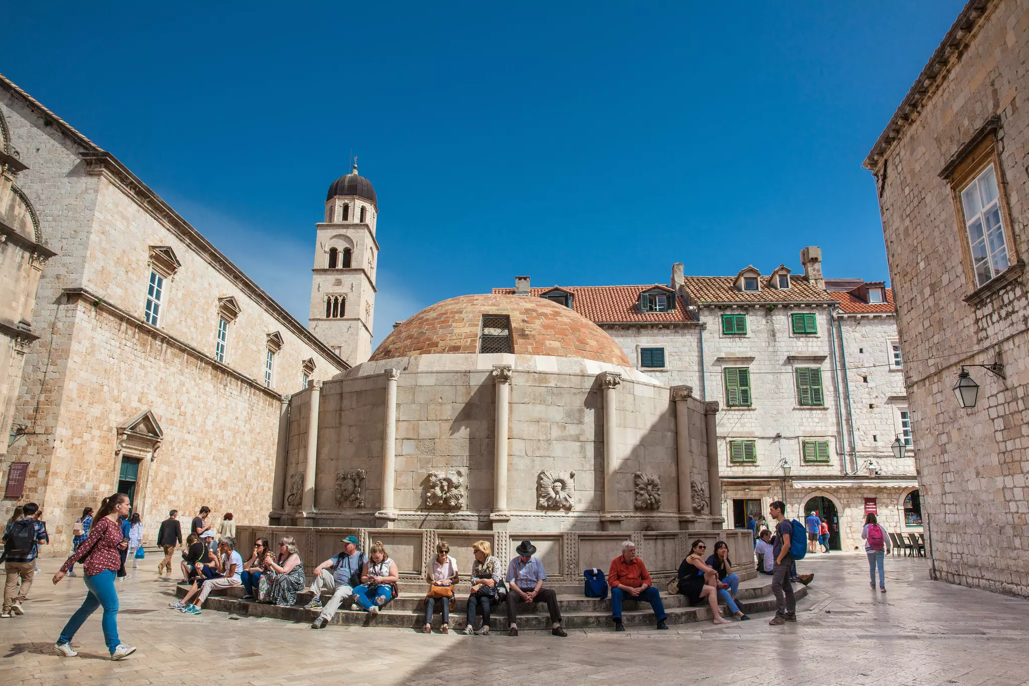 Tourists sat around a large rotunda on a sunny day, with a bell tower rising above them and the old town.