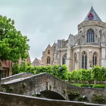 Onze-Lieve-Vrouwekerk in Bruges, its buttressed stone apse and slate spire seen over a small arched brick bridge and trees