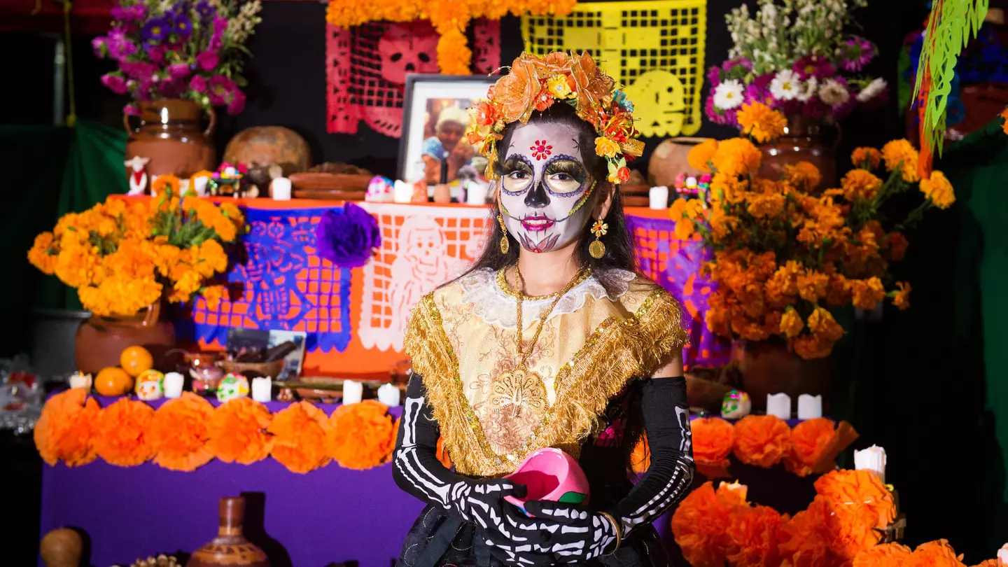 Girl in skeleton costume and face paint stands in front of an altar for the Day of the Dead festival in Mexico