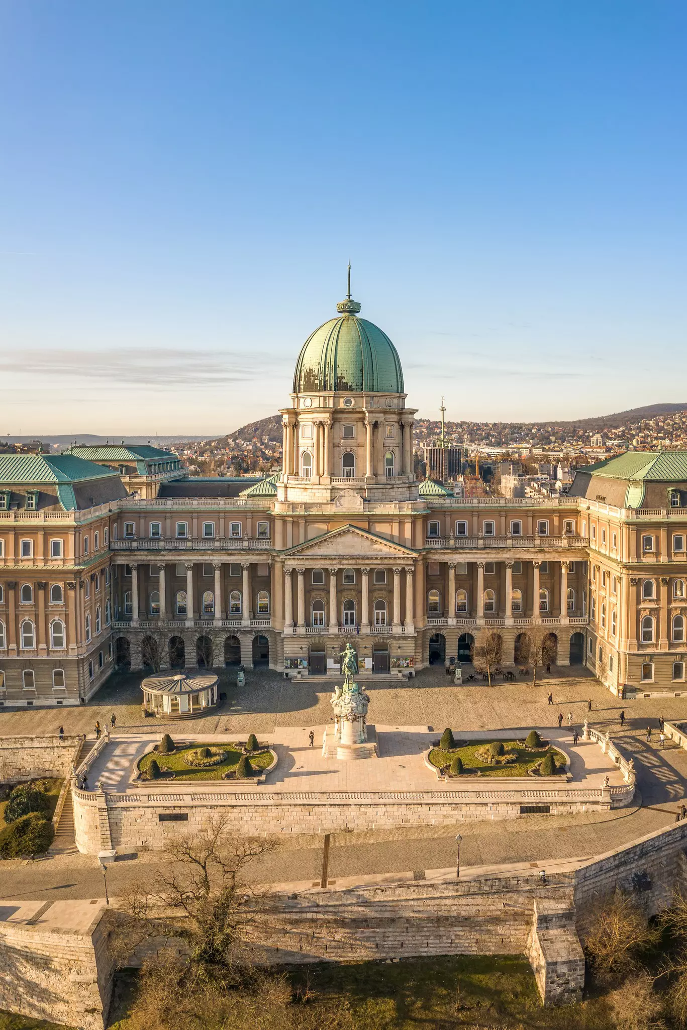 An aerial view of Budapest's Castle District on a sunny day. The old stone buildings are surrounded by pockets of greenery.