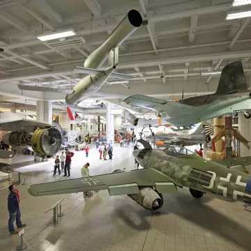 A shot taken from above looking down into a massive museum room furnished with airplanes and other vehicles. People are moving around between the exhibits