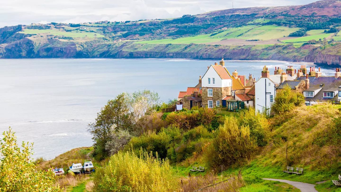 A small path leads to a fishing community with historic white buildings with orange roofs. The town sits on the edge of a bay with rolling green hills in the background. England.