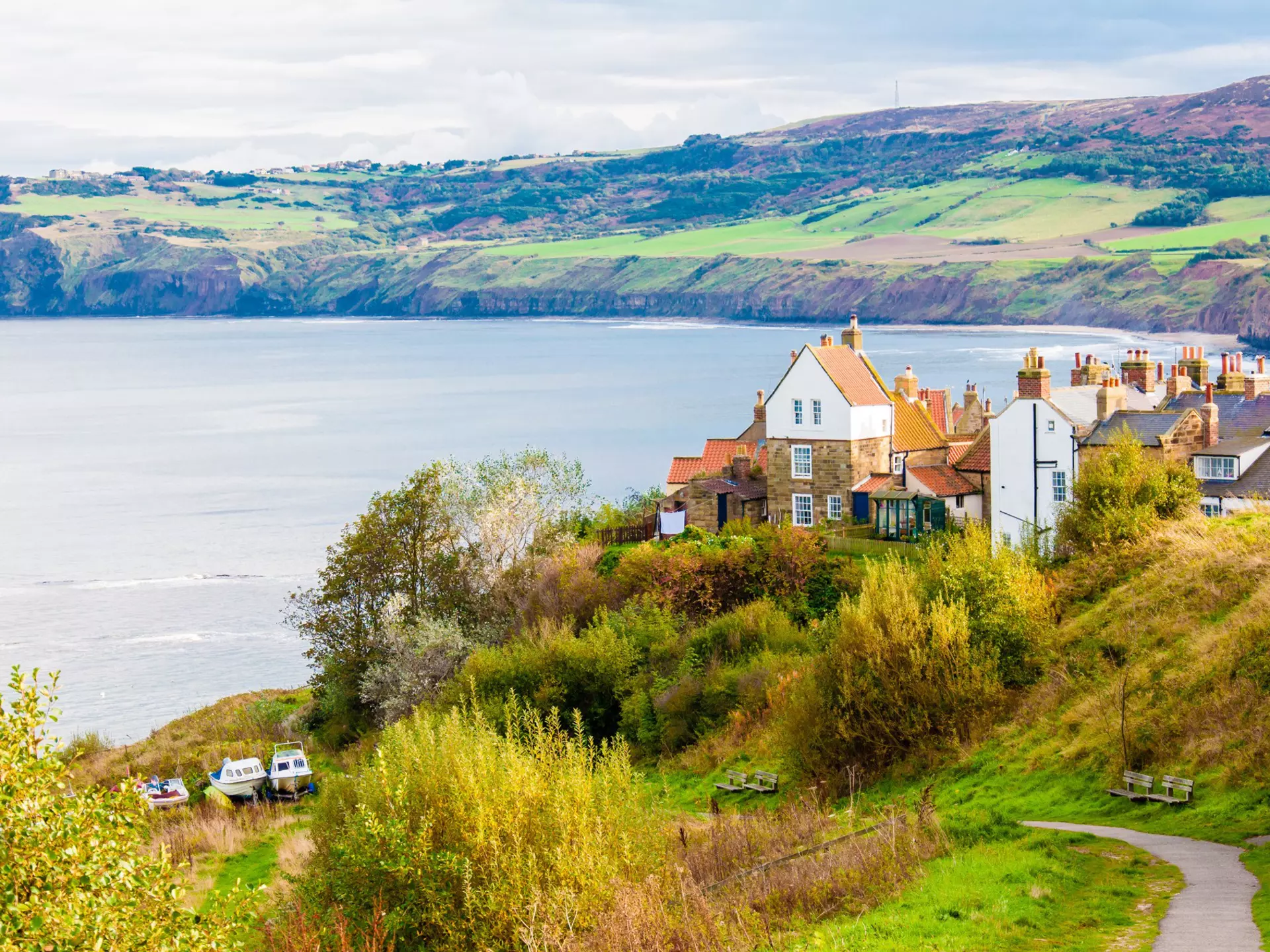 A small path leads to a fishing community with historic white buildings with orange roofs. The town sits on the edge of a bay with rolling green hills in the background. England.