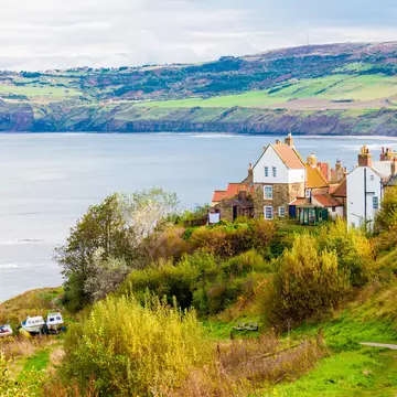A small path leads to a fishing community with historic white buildings with orange roofs. The town sits on the edge of a bay with rolling green hills in the background. England.
