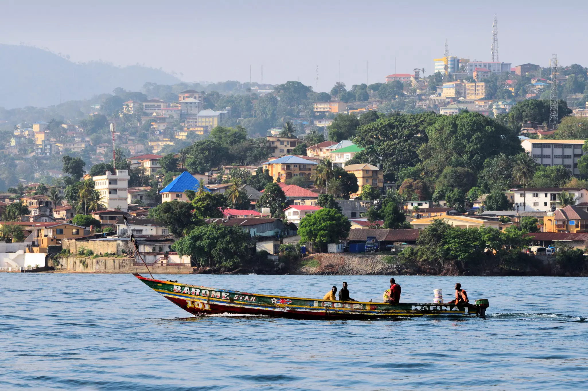 People ride a long, brightly painted traditional fishing boat through a bay past a city, where buildings rise on a hill on the shore.
