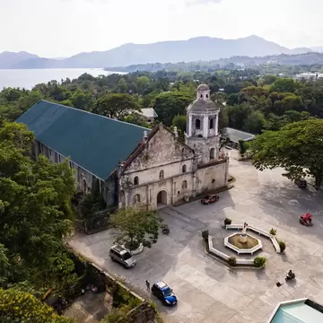 Old stone church with a bell tower and green roof beside a paved plaza, surrounded by trees with the sea and hills behind.