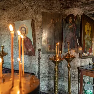 Painted Orthodox icons hang on a rough carved stone wall beside brass candlestands holding thin lit candles.
