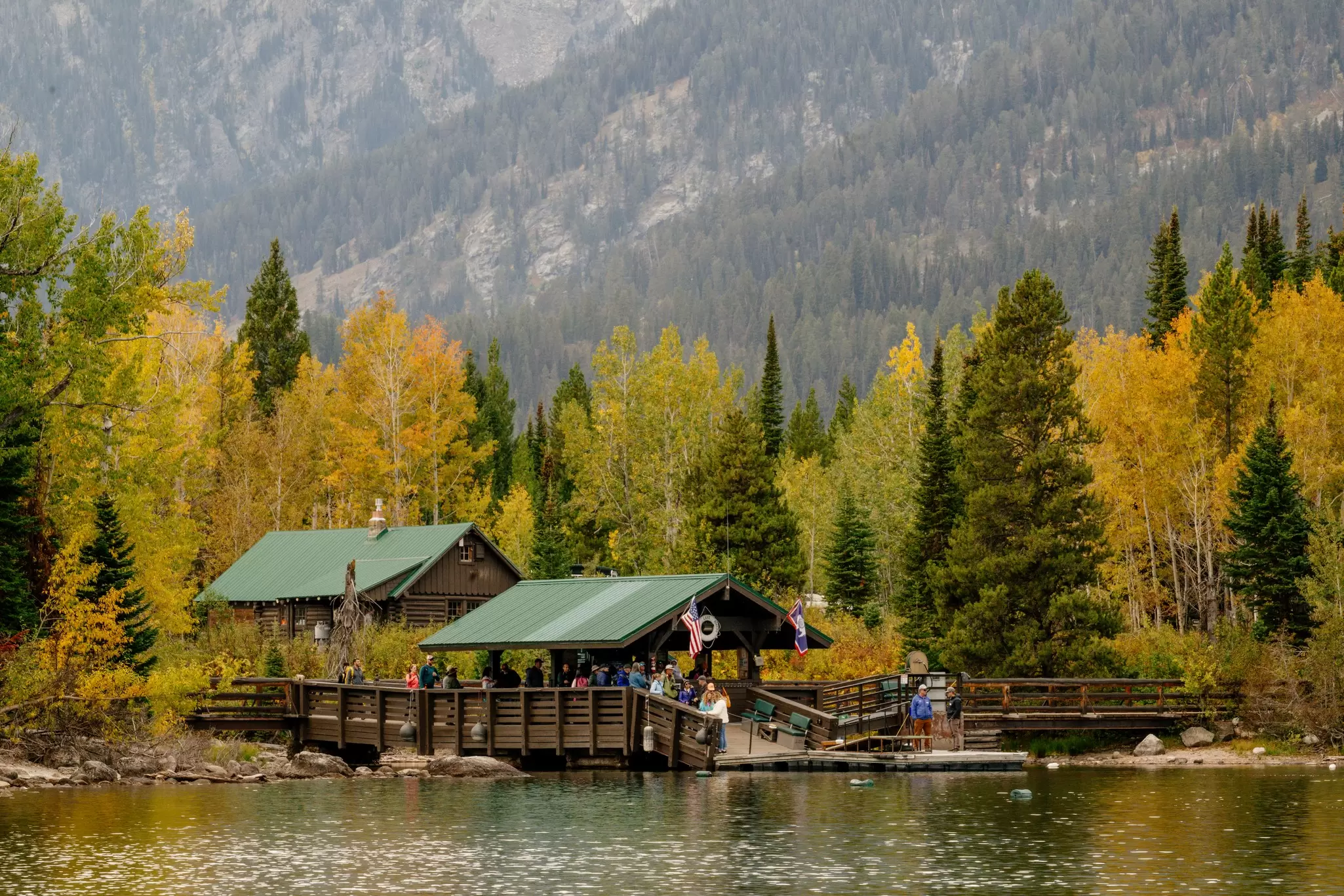 A group of people walk onto a dock sitting on a lake. The dock and building behind it are brown with green roofing, and there is bright yellow fall foliage and mountains in the background.