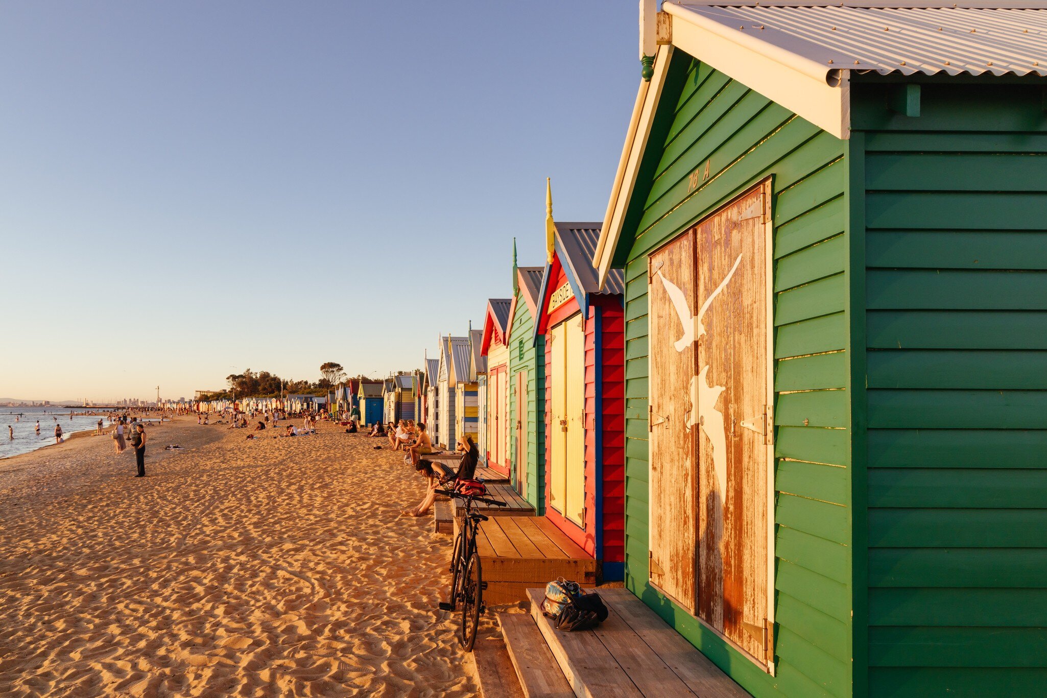 People sitting along the colorful Brighton Beach bathing boxes at Denby St beach and golden sand. 