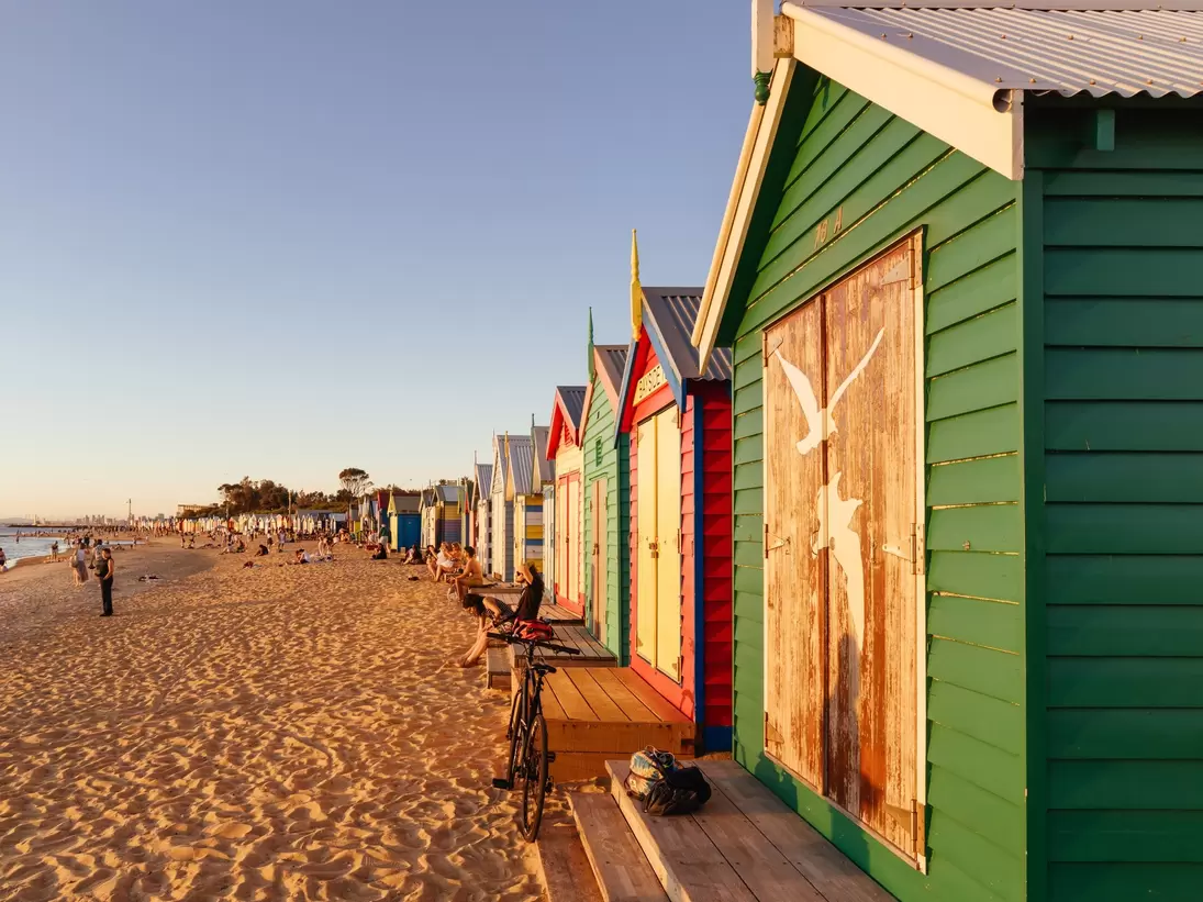 People sitting along the colorful Brighton Beach bathing boxes at Denby St beach and golden sand. 